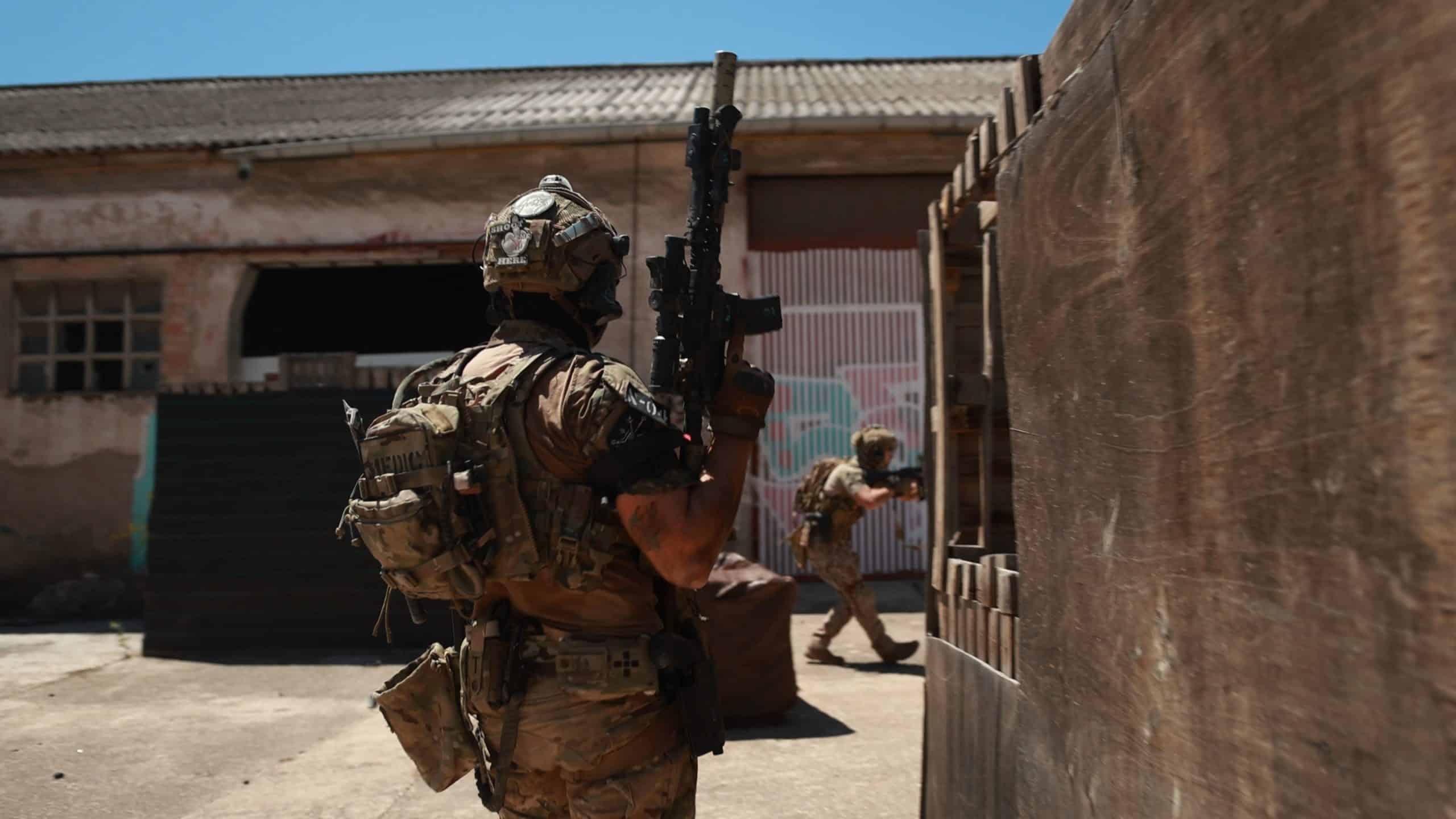 Dos jugadores de airsoft avanzando junto a una nave en un campo urbano